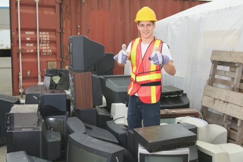 A man wearing a yellow safety helmet, high-visibility vest with orange and yellow stripes, and gloves is standing outdoors at a rubbish removal site in front of a collection of old, discarded electronic devices including large CRT televisions, monitors, and computer towers with black and gray casings, some with visible screens and cables. The background features a red shipping container, a wooden pallet, and a partially visible white tent or cover, suggesting an industrial or disposal environment. The scene is well-lit by natural daylight, highlighting the textures and colors of the electronics and safety gear, and reflects a professional approach to domestic or commercial rubbish clearance in London or nearby areas.