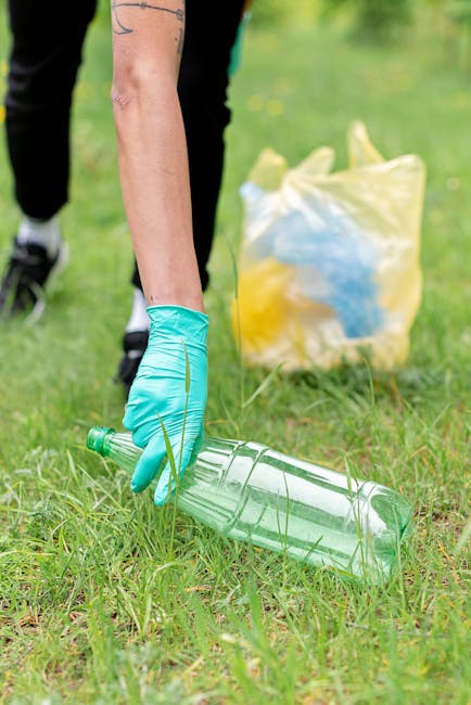 A waste collection worker wearing a high-visibility yellow and orange jacket is operating a red rubbish collection truck, positioned on a street with partially visible roadside grass and sidewalk. The
