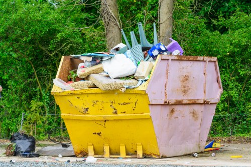 A large, rectangular skip bin placed outdoors on a gravel surface, filled with various types of rubbish including plastic laundry baskets, fabric and paper waste, wooden and cardboard materials, and other household debris. The skip features a yellow lower section and a pinkish upper section with visible dirt and rust markings. It is positioned adjacent to a green area with trees and dense foliage, suggesting a suburban or park-like setting. Nearby, a black plastic bag and small scattered debris are present on the ground. The image depicts a typical scene of waste clearance, perhaps in the context of house or garden rubbish removal services in Crouch End, London, provided by Crouch End House Clearance.