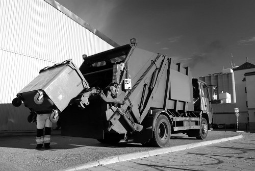 A commercial refuse collection vehicle, parked on a paved surface in an industrial or urban area, with its large rear container raised to unload waste. The truck has a robust metal body with a textured finish, typical of waste management vehicles, painted in a dark tone. Mounted on the back are hydraulic arms and mechanisms for lifting and tipping rubbish containers, with some visible components appearing metallic and slightly reflective. To the left, a person wearing work trousers and safety gloves is positioning a large waste bin, which has a smooth, rectangular shape and is made of plastic or metal. The bin is slightly tilted, suggesting it is being emptied into the truck's container. In the background, there is a tall wall or building with a corrugated metal surface, and the sky appears partly cloudy with sunlight casting shadows, indicating daytime. The scene reflects a typical rubbish collection operation, consistent with the services offered by Crouch End House Clearance and similar providers in the Crouch End area, N8 postcode.