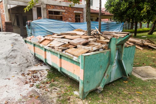A large, rectangular green metal skip filled with a variety of wooden planks, panels, and debris, positioned on a patch of grass near a residential area. The wood pieces are untreated, irregular in size, and show signs of weathering with rough textures and light brown to dark brown tones. The skip's sides are reinforced with orange reflective strips, and the lid is open, revealing the clutter inside. Behind the skip, there are two blue tarpaulins secured with ropes, covering construction materials or additional debris, with a partially built brick structure visible in the background. To the right, a tree with green foliage provides shade, and the setting appears to be a suburban street with residential buildings and a paved sidewalk nearby. The scene is captured during daylight, with natural light illuminating the outdoor environment, indicative of a typical rubbish removal task in the Crouch End or nearby London area, managed by Crouch End House Clearance.