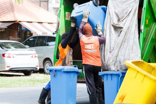Two waste collection operatives wearing high-visibility orange safety vests and gloves are positioned beside a green rubbish truck on a residential street, with trees and parked cars visible in the background. One worker is reaching into the truck to handle a large blue plastic bin, while the other appears to be assisting or observing, with a blue bin placed nearby on the pavement. A yellow bin is also partially visible in the foreground, next to the blue bins. The scene takes place during daylight, with natural light illuminating the area. The street setting suggests a house clearance or rubbish removal service in Crouch End, highlighting the practical process of waste collection and disposal for local clients managed by Crouch End House Clearance. The materials include plastic waste containers, rubber gloves, and the metallic surface of the waste truck, with textures ranging from smooth plastics to the reflective metal and fabric of the workers' clothing. The background includes residential elements such as parked cars and buildings, framing a typical urban scene focused on responsible rubbish disposal in London.