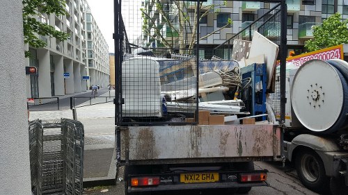 A flatbed truck with an open metal cage at the rear, parked on a city street corner, is loaded with various types of household rubbish including white PVC window frames, wooden panels, a large white plastic container, and miscellaneous debris. The truck's surface shows signs of rust and wear, and the cargo spills slightly over the sides of the bed. The background features modern residential buildings with balconies, a traffic light, and a crosswalk, indicating an urban environment consistent with areas around Crouch End. Adjacent to the truck on the pavement are metal shopping carts, suggesting the removal or clearance of waste or unwanted items typical of house clearance services. The scene is captured during daylight hours, with natural light illuminating the surroundings, emphasizing the textures and materials of the discarded items and the vehicle used for rubbish removal.